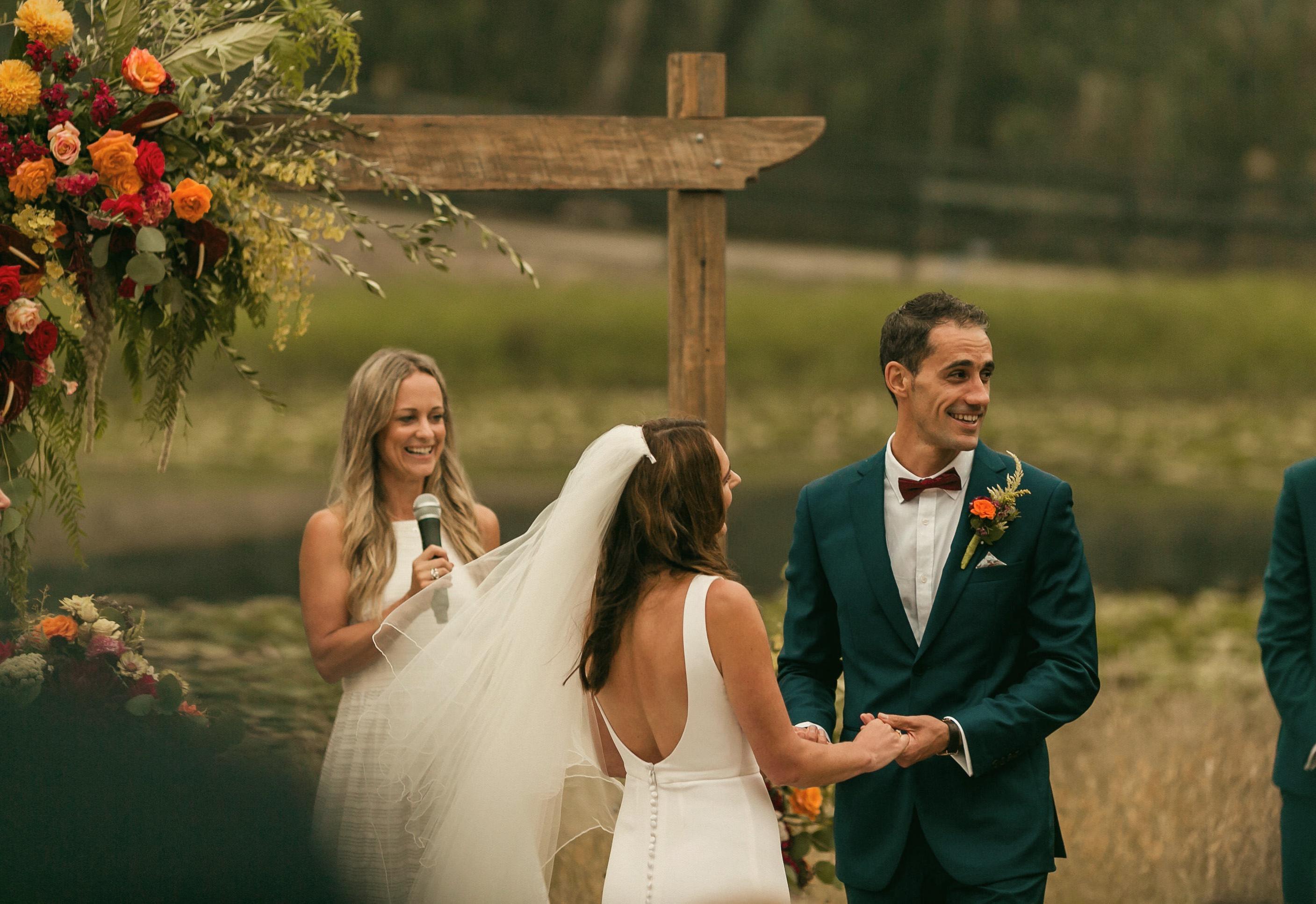 A couple surrounded by guests at a beachside ceremony, mid-laugh during their vows.